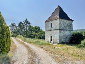 Una torre de piedra con techo a dos aguas, un sendero de grava que la conduce y follaje circundante.