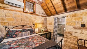 Una habitación rústica con pared de piedra, vigas de madera en el techo, una cama con ropa de cama floral, una mesita de noche con lámpara y un pequeño ventanal con cortina.