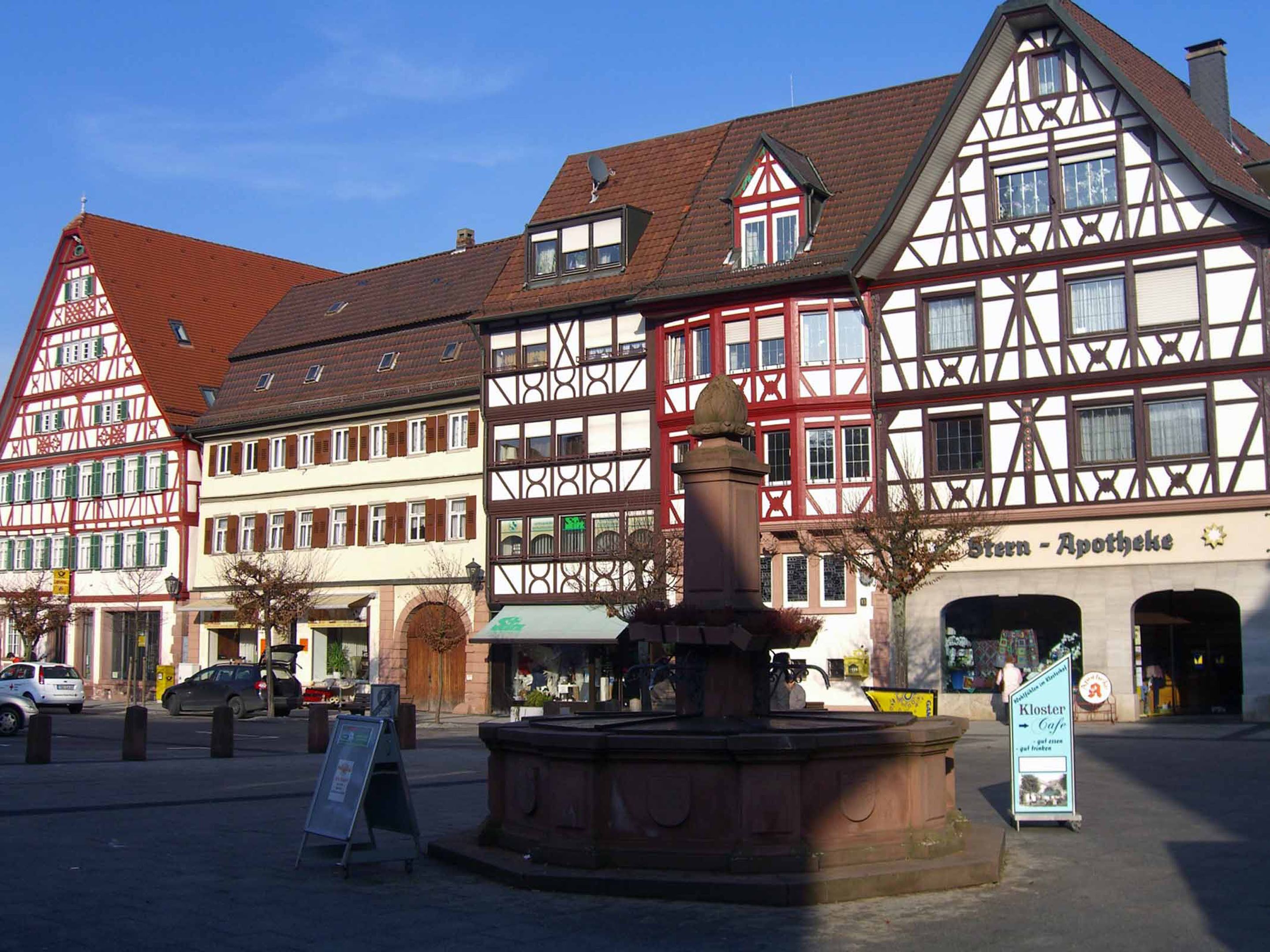 A traditional European town square with half-timbered buildings, a fountain, and a sign for 'Stern - Apotheke'.