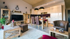 Una sala de estar moderna con techo de madera, televisor, zona de comedor con sillas y una cocina al fondo.