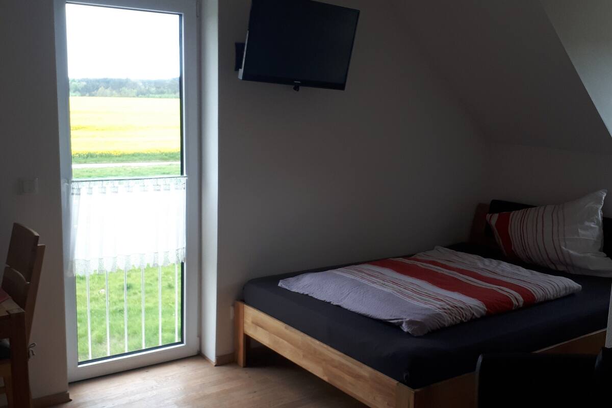 Ein Schlafzimmer mit Bett, Stuhl, Fernseher und einem Balkon mit Blick auf ein Feld.