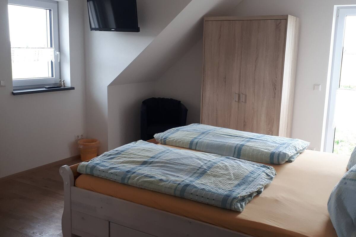 Ein Schlafzimmer mit schrägen Wänden, einem Holzschrank, einem Bett mit Karo-Decke und einem Fenster mit Blick auf Grünflächen.