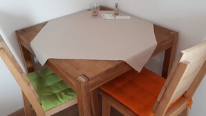 Una pequeña mesa de madera con mantel blanco, dos sillas con cojines verdes y anaranjados, y una pequeña vela y figurita sobre la mesa.