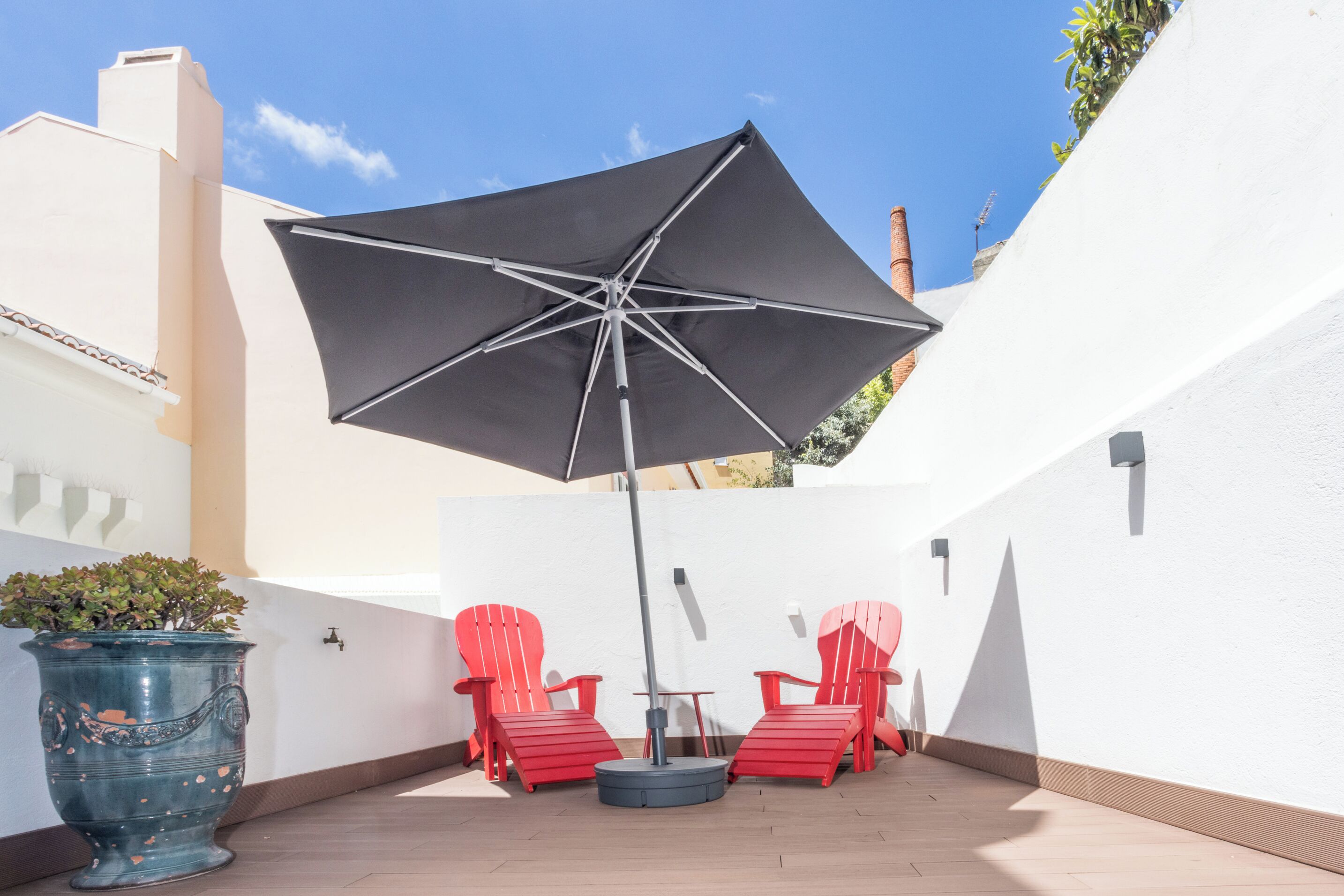 En terrasse med en stor sort parasol, to røde stole og en potteplante.