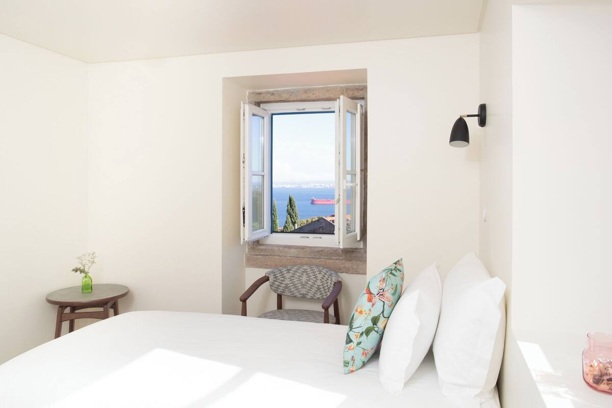 Un dormitorio con cama, una silla, una mesita de noche con un florero y una ventana que ofrece vistas abiertas al paisaje.
