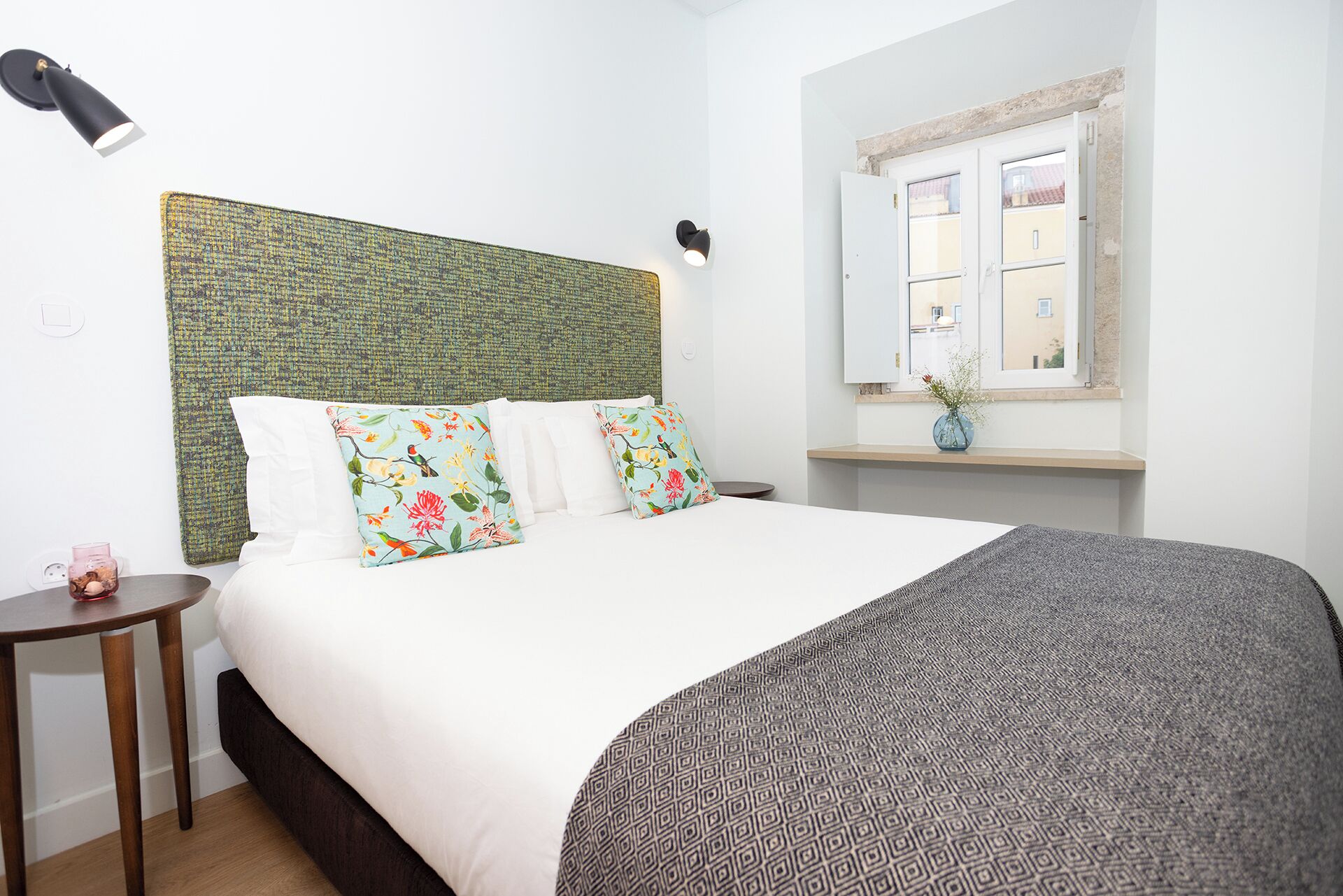 A neatly made bed with floral pillows, a textured green headboard, a bedside table with a vase, and a window with a view of buildings.