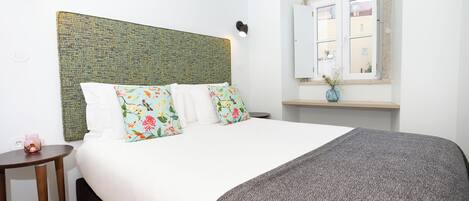 A neatly made bed with floral pillows, a textured green headboard, a bedside table with a vase, and a window with a view of buildings.