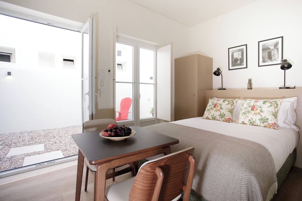 Un dormitorio con cama, mesita de noche, silla, ventana y puerta.