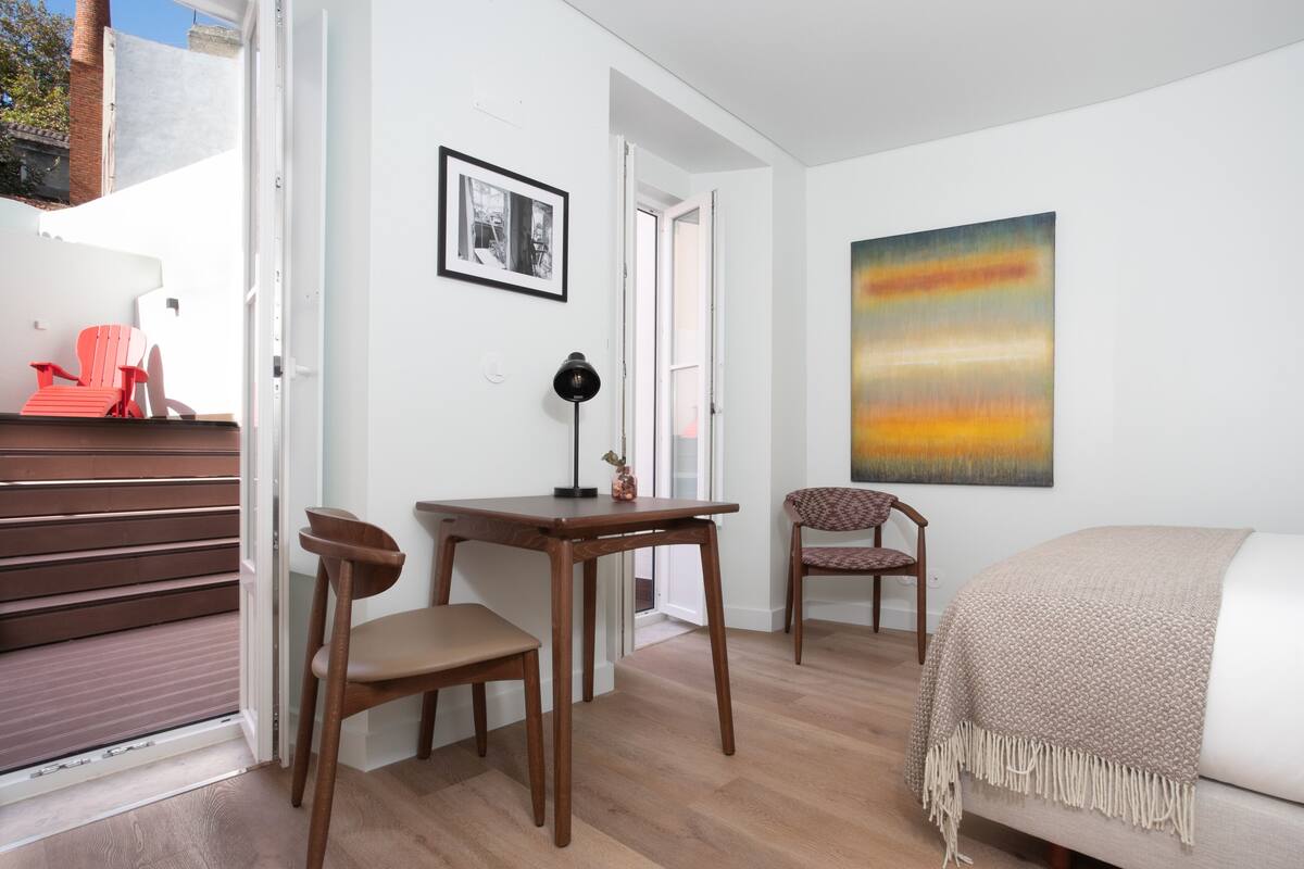 Una habitación con cama, escritorio, dos sillas, lámpara y un cuadro en la pared.