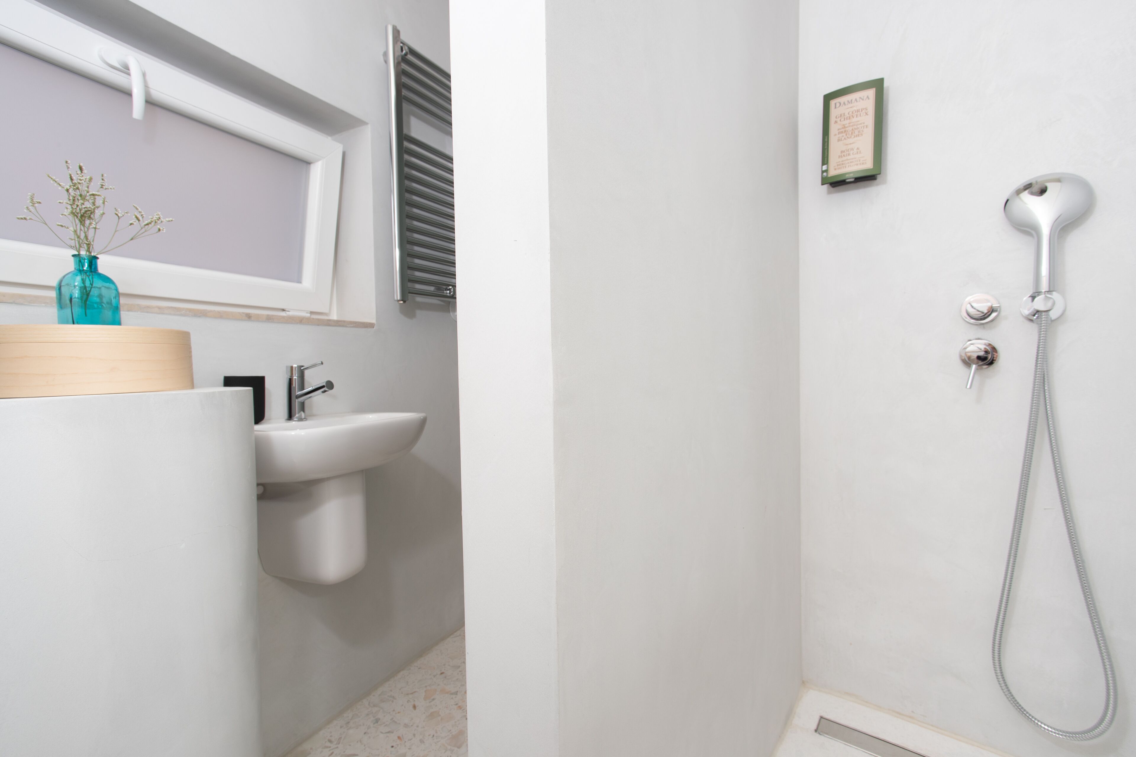 Ein Badezimmer mit einem weißen Waschbecken, einem Fenster mit Jalousien, einer Dusche mit Duschbrausehandstück und einem gerahmten Zertifikat an der Wand.