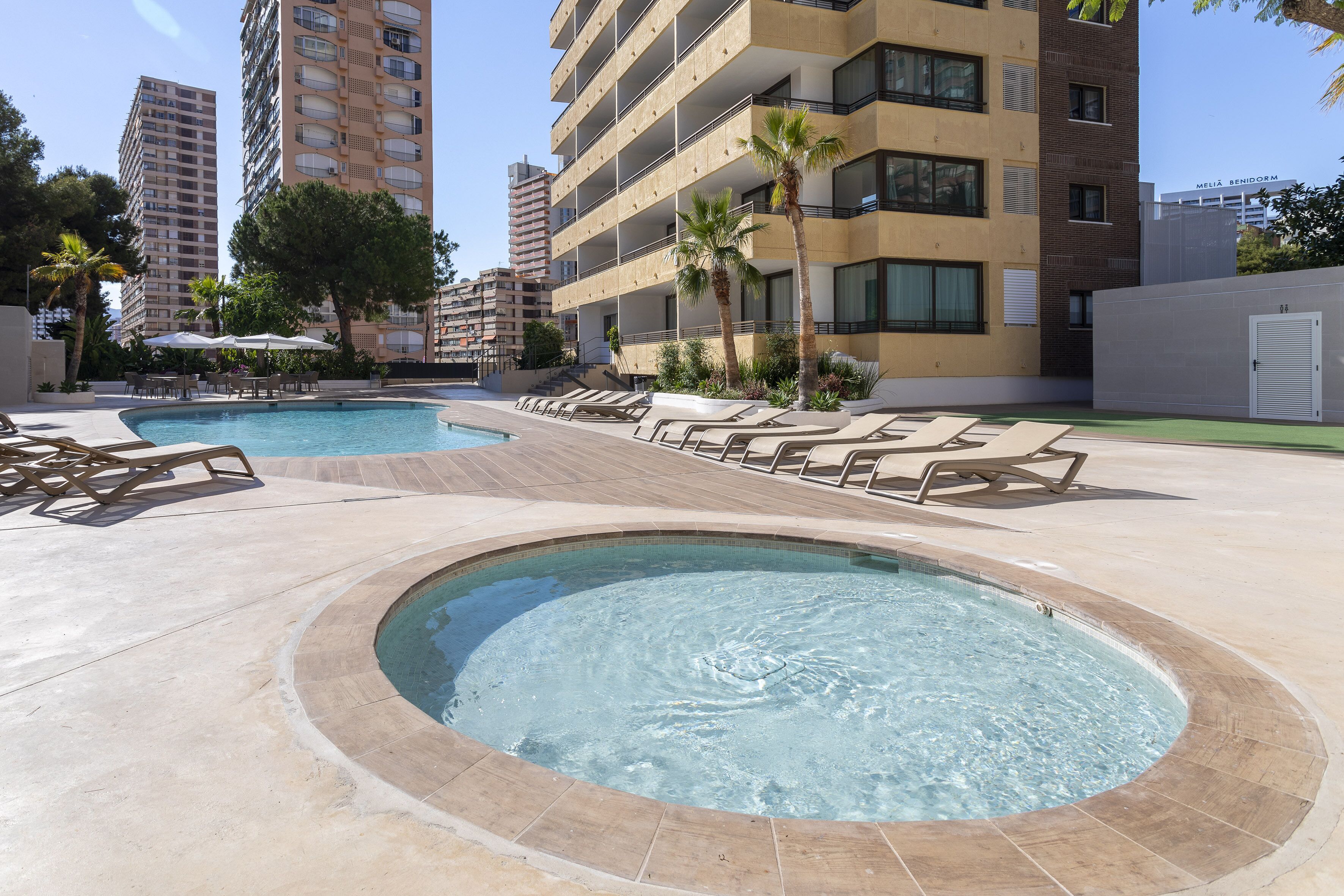 Zona de piscina con sillones de descanso y jacuzzi, rodeada de edificios y palmeras.