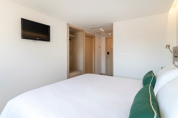 Ein Hotelzimmer mit einem Bett, einem an der Wand befestigten Fernseher, einem Kleiderschrank und einer Tür zu einem weiteren Raum.