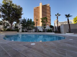 A swimming pool with a tiled deck, surrounded by greenery and a tall building in the background.