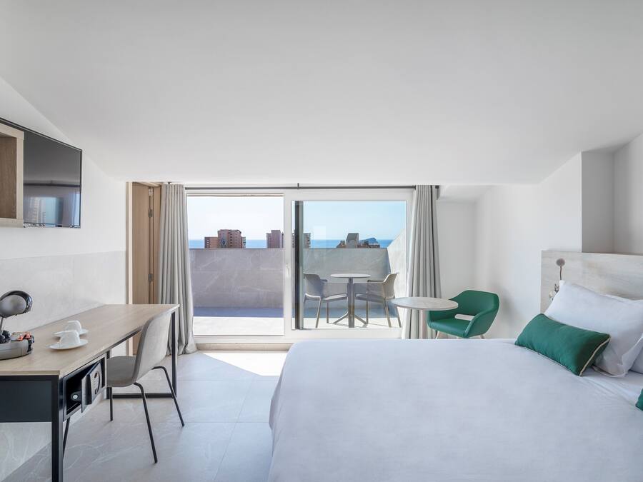 A hotel room with a bed, a desk, a chair, a small table, and a balcony with a view of the city.