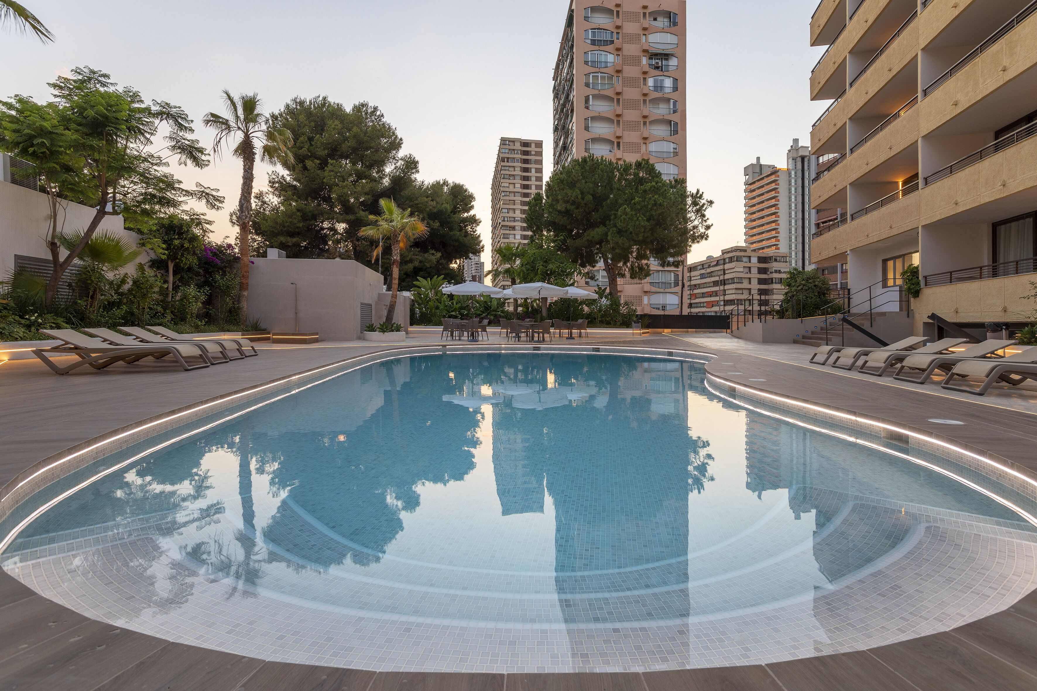 A swimming pool with lounge chairs and umbrellas, surrounded by tall buildings and trees.