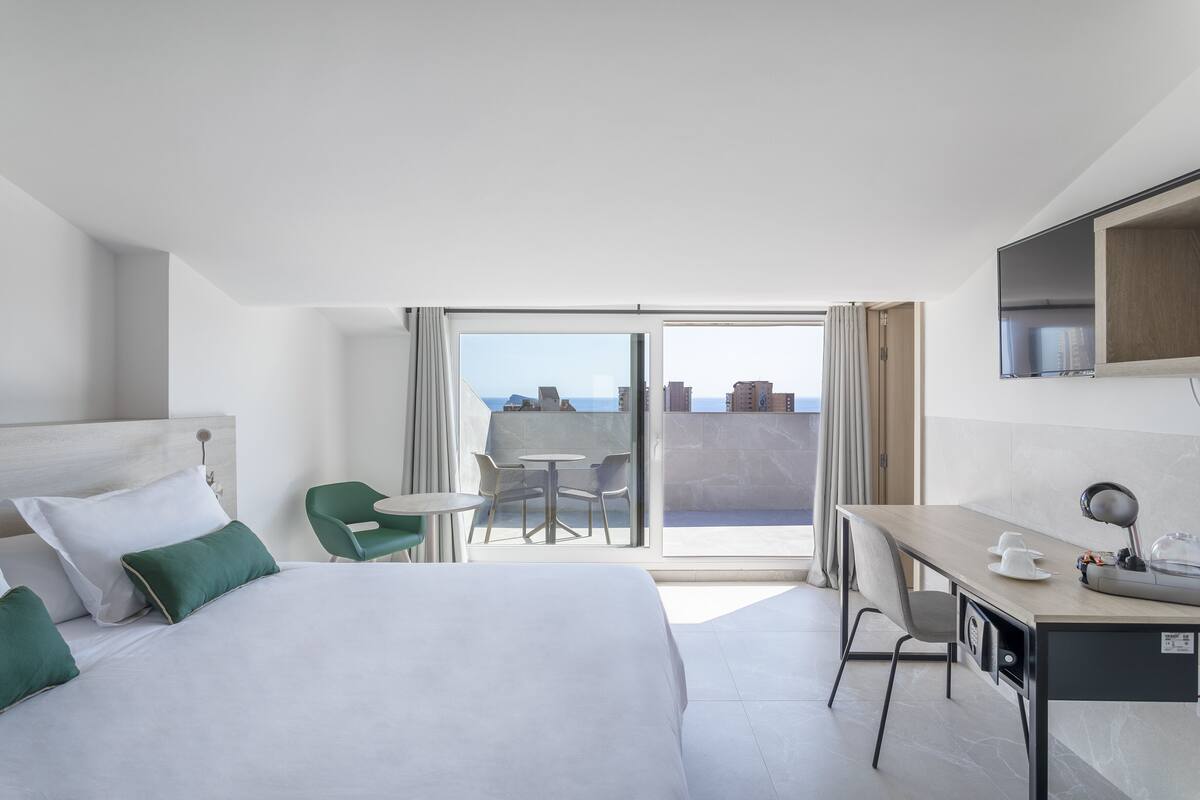 Habitación de hotel con cama, escritorio, silla, mesa y balcón con vistas.