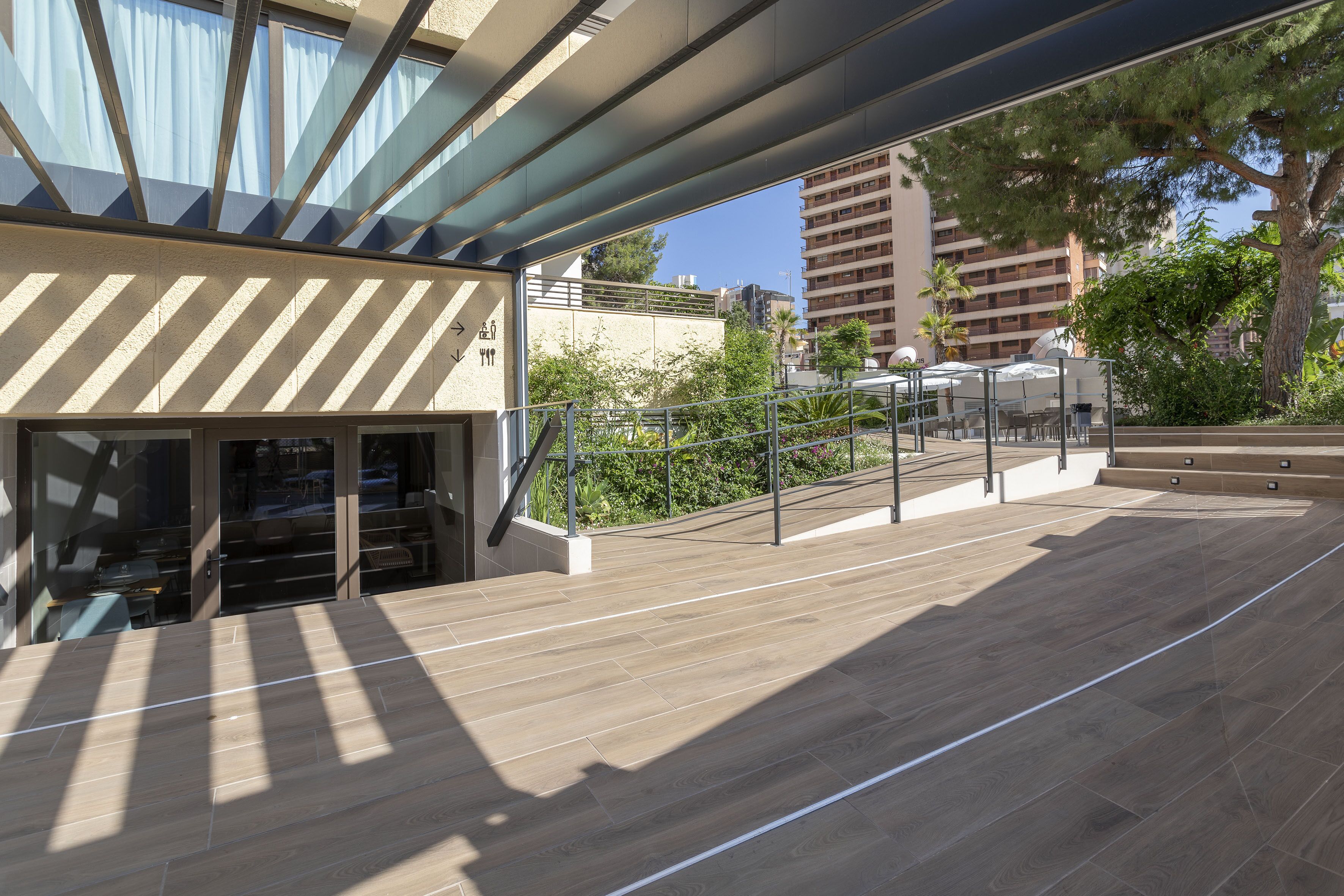 Un espacio exterior moderno con terraza de madera, puerta de entrada de vidrio y vistas a los edificios y la vegetación circundantes.