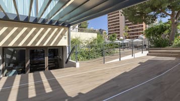Un espace extérieur moderne doté d’une terrasse en bois, d’une porte d’entrée en verre et offrant une vue sur les bâtiments et la végétation environnants.