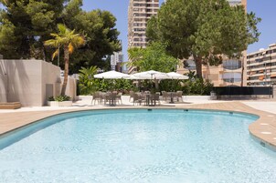 A swimming pool with lounge chairs and umbrellas, surrounded by trees and modern buildings.