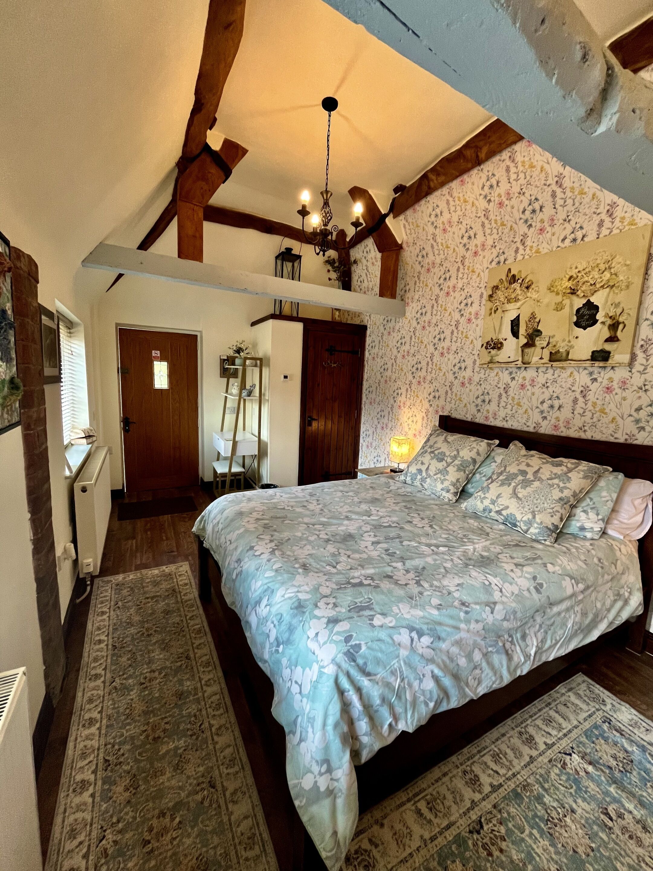 A bedroom with a large bed, wooden beams, a chandelier, a floral wallpaper, and a small table with a lamp.