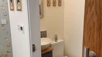 Un baño con inodoro blanco, tocador de madera con un lavamanos grande y zona de ducha. Hay tres cuadros enmarcados en la pared sobre el tocador.