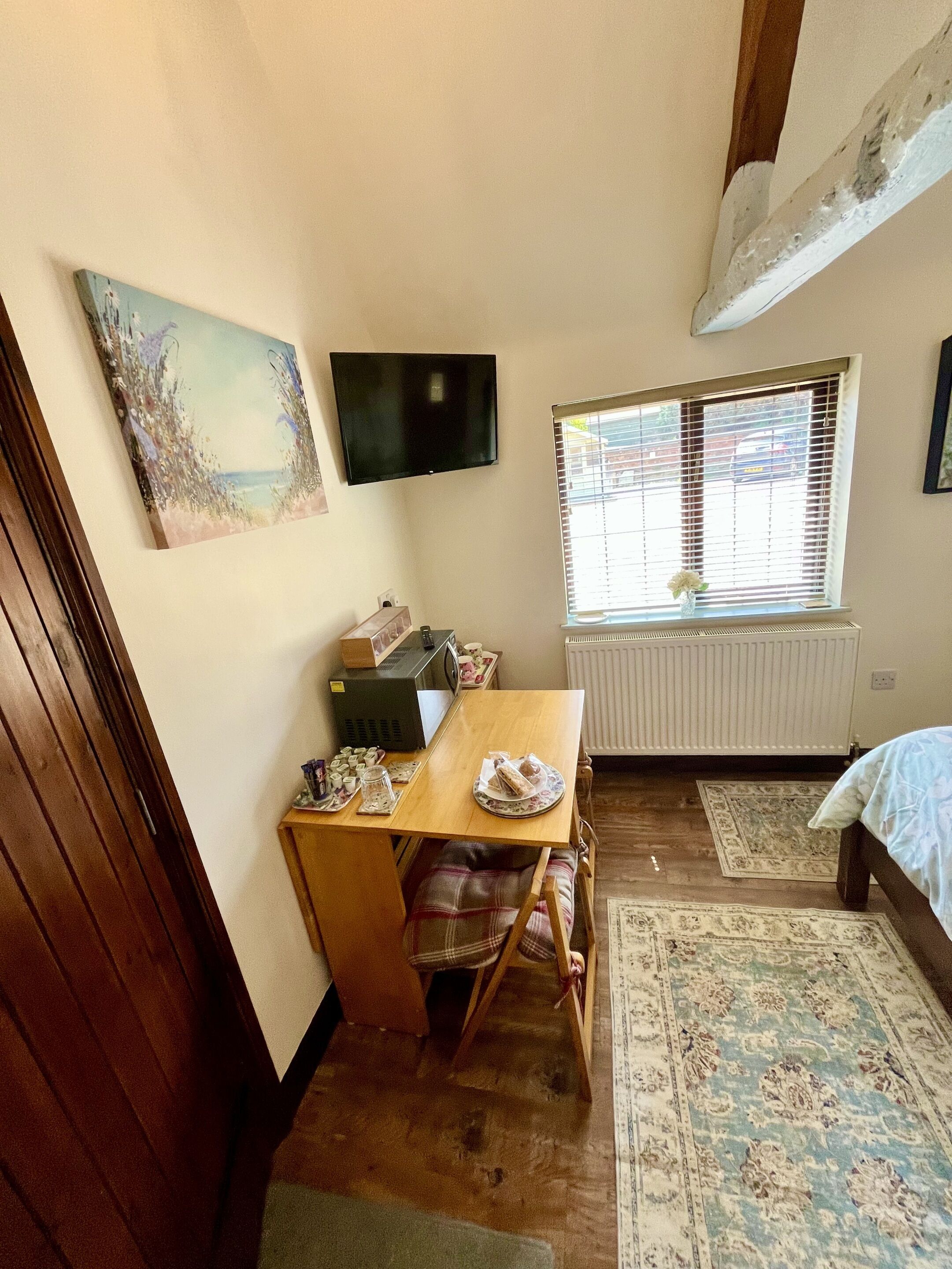 A small, cozy room with a wooden table, a TV mounted on the wall, a window with blinds, and a radiator.