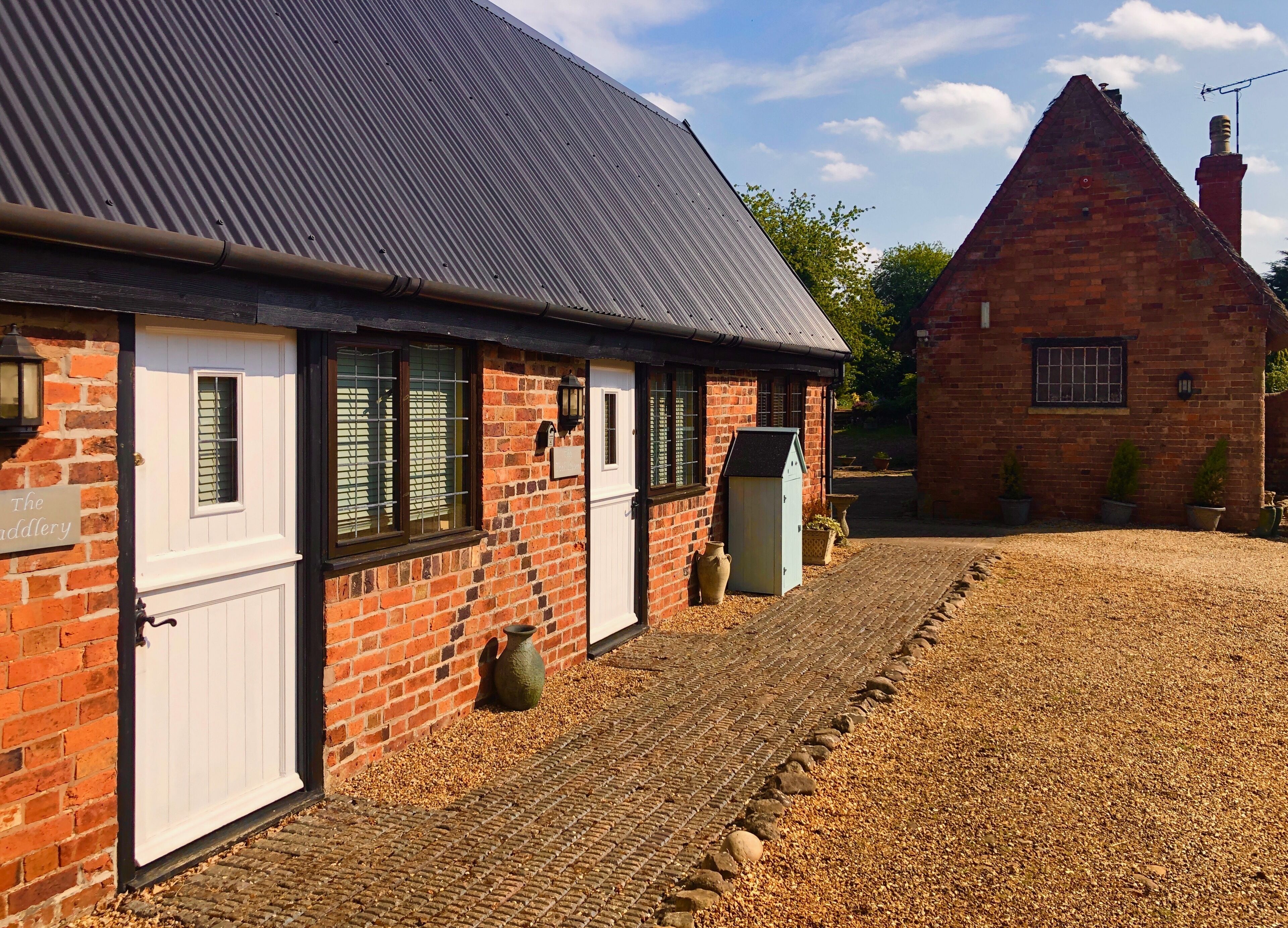 A brick building with a white door and windows, a gravel driveway, and a smaller brick building in the background.