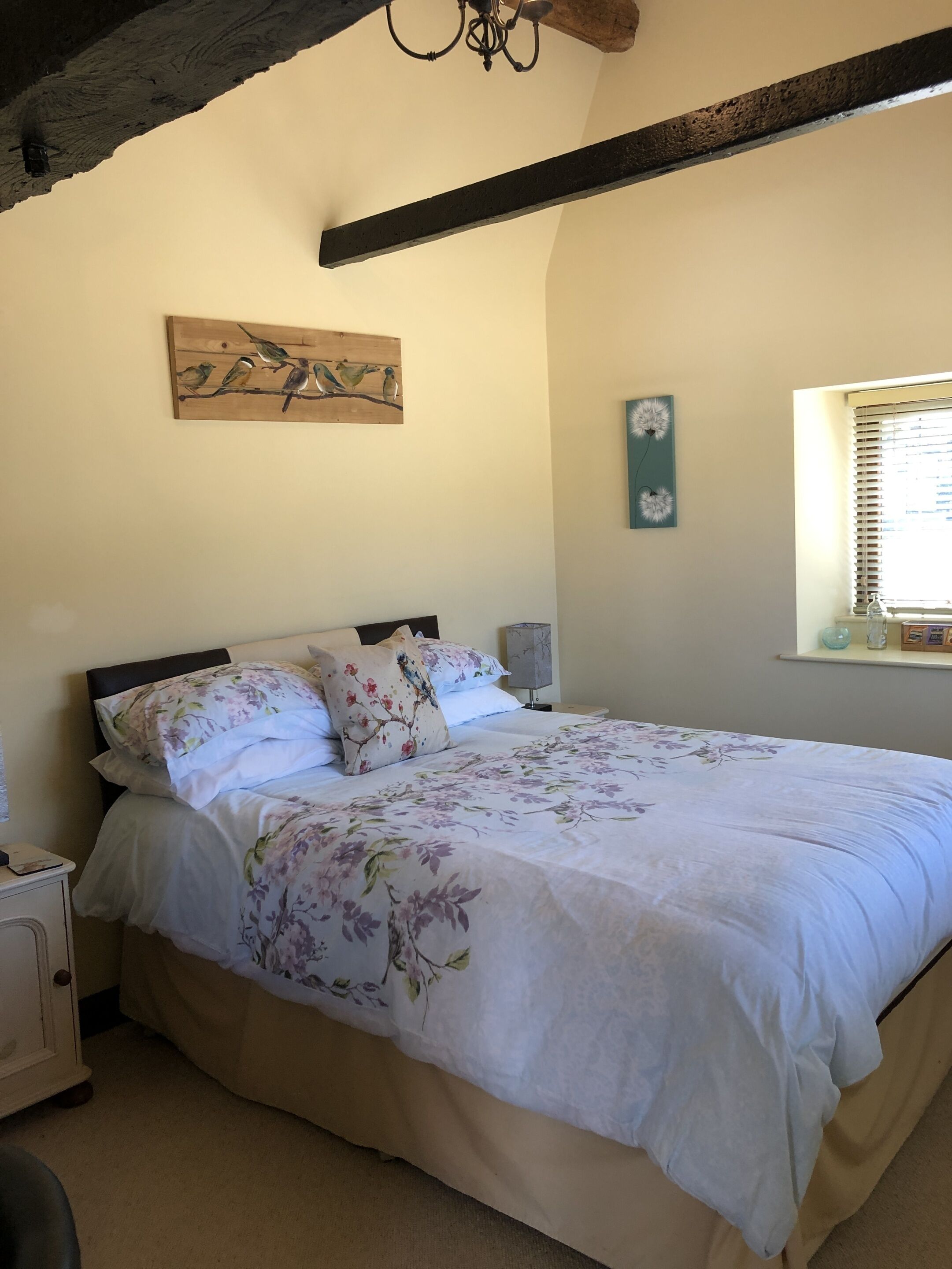 A bedroom with a large bed, a wooden ceiling beam, a wall-mounted artwork of birds, a window with blinds, and a bedside table with a lamp.