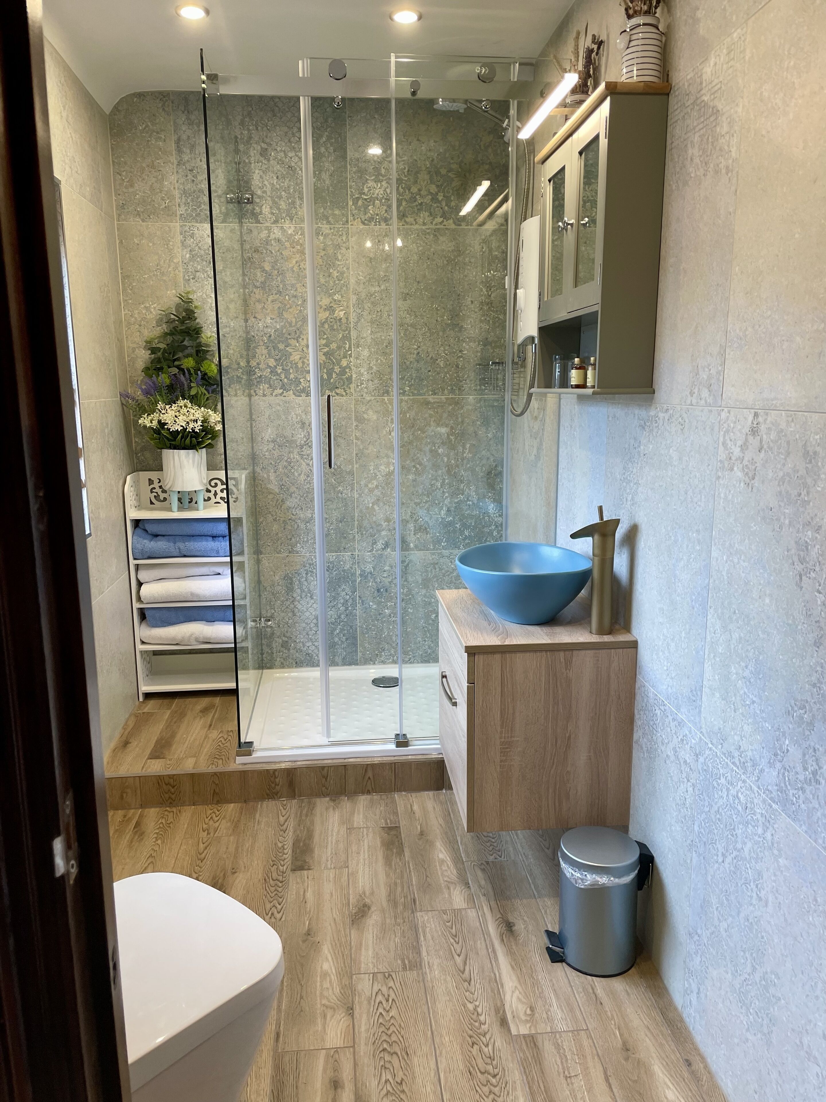 A modern bathroom with a glass-enclosed shower, a wooden vanity with a blue basin, and a white toilet.