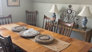 A dining room with a wooden table, woven placemats, and chairs. There are two wall-mounted lamps and a framed picture on the wall.