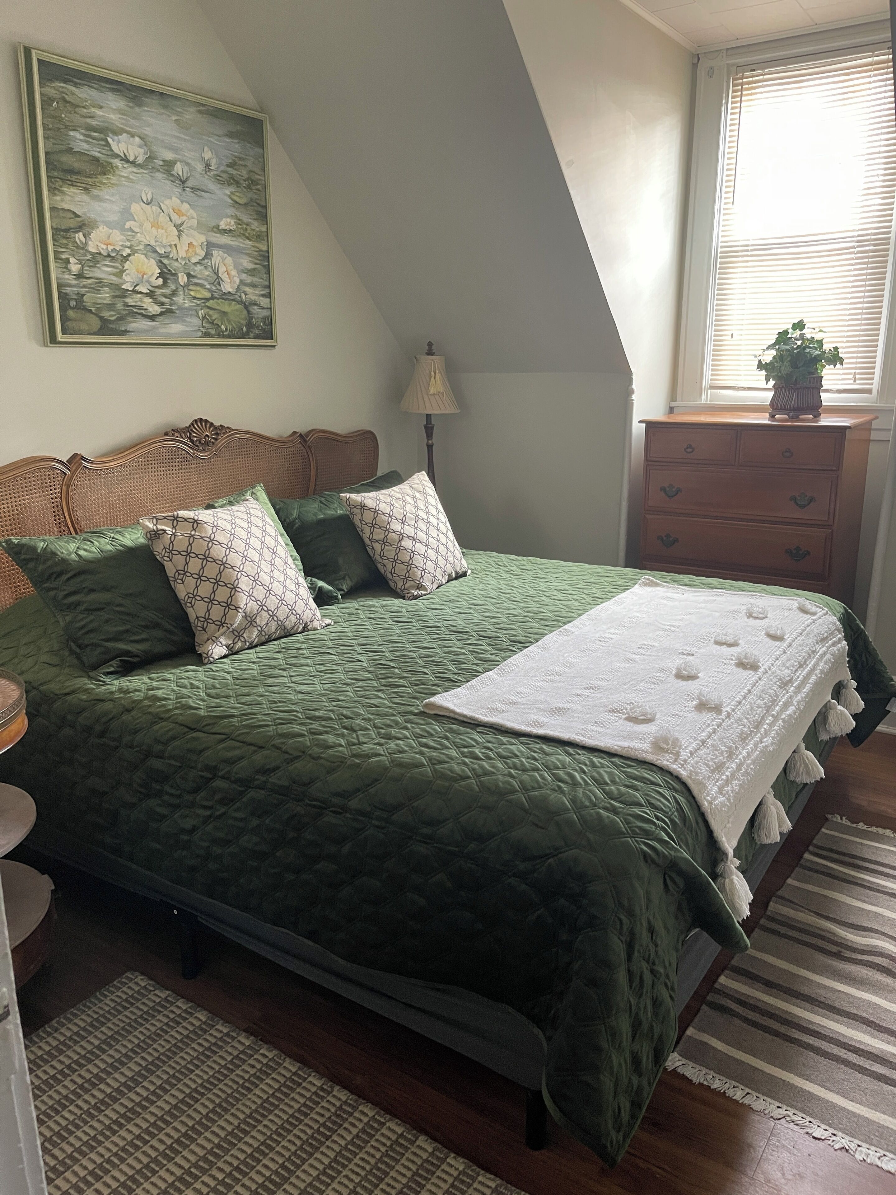 A bedroom with a green quilted bedspread, a wooden headboard, a bedside lamp, a dresser, and a window with blinds.