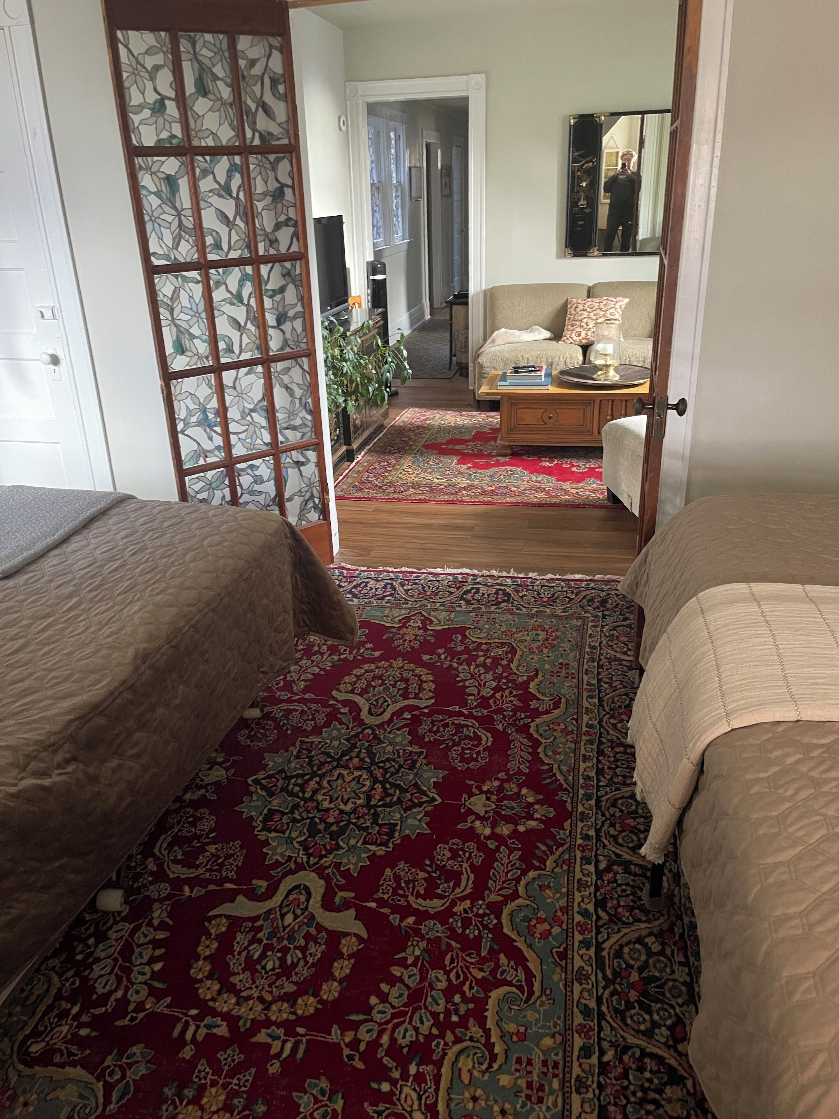 Habitación con dos camas, alfombra roja con estampado, puerta de madera con panel decorativo y un pasillo que conduce a otra habitación.