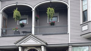 A two-story house with a front porch, a central entrance with double doors, and a balcony with hanging flower baskets.