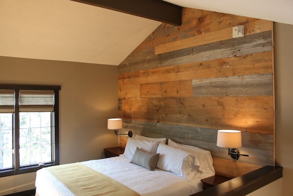 Quarto com teto inclinado, parede revestida de madeira, duas mesas de cabeceira com luminárias, cama com roupa de cama branca e janela com persianas.