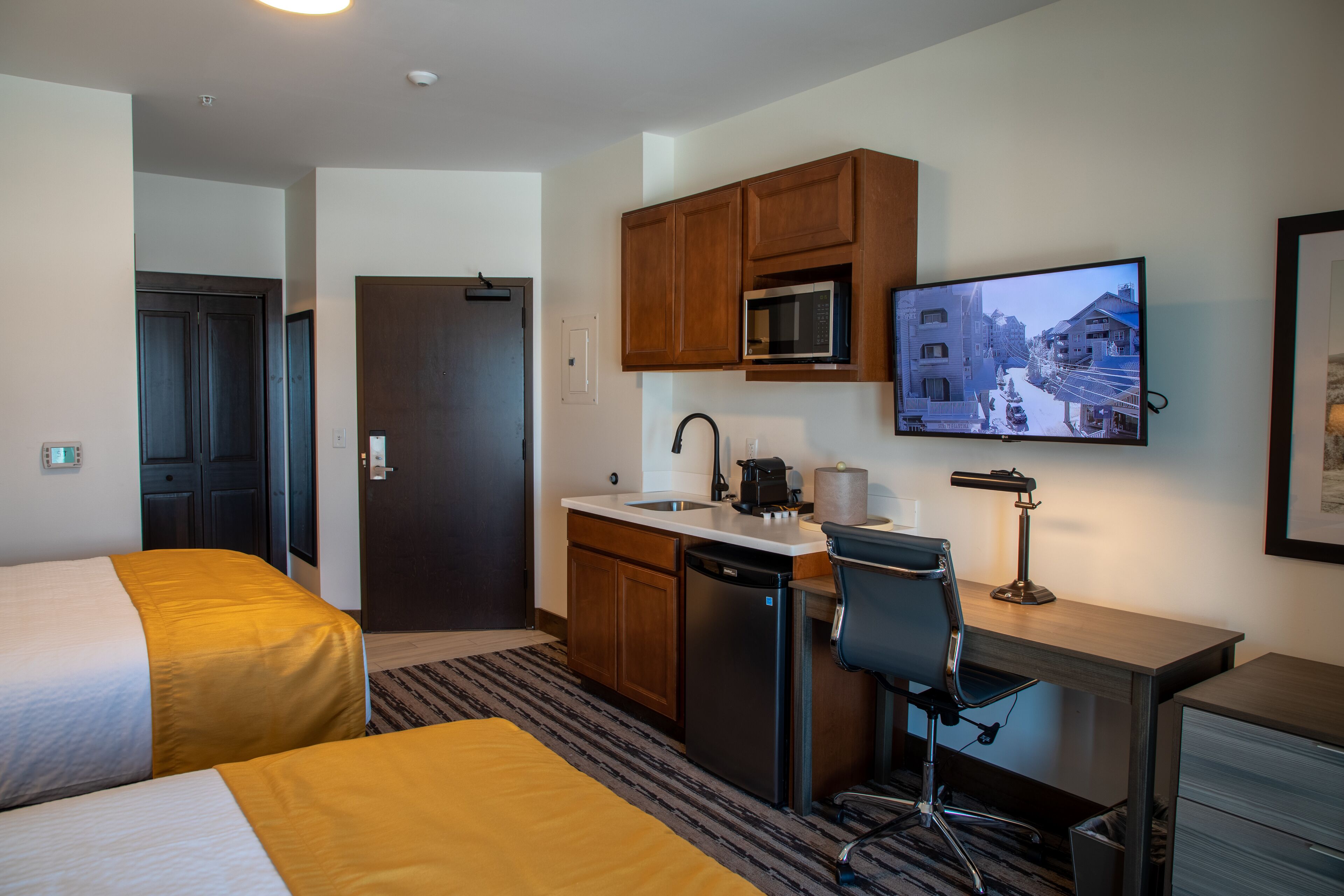A hotel room with two beds, a kitchenette, a desk with a chair, a television, and a door.