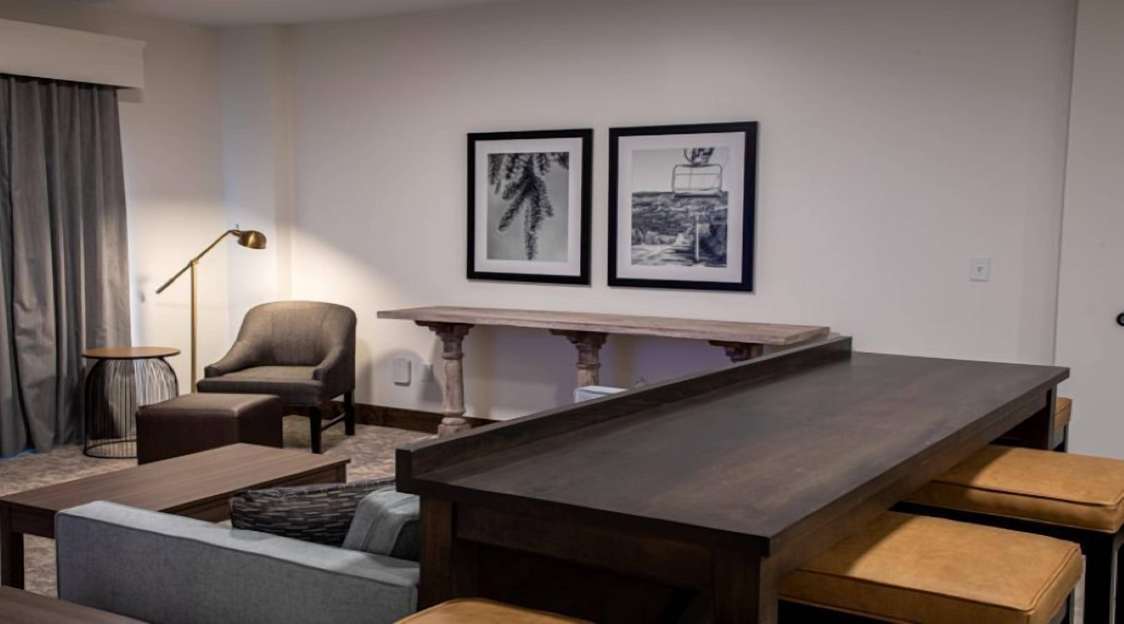 Un salon moderne avec une longue table en bois, un fauteuil gris, une petite table ronde et un canapé. Deux tableaux encadrés sont accrochés au mur.
