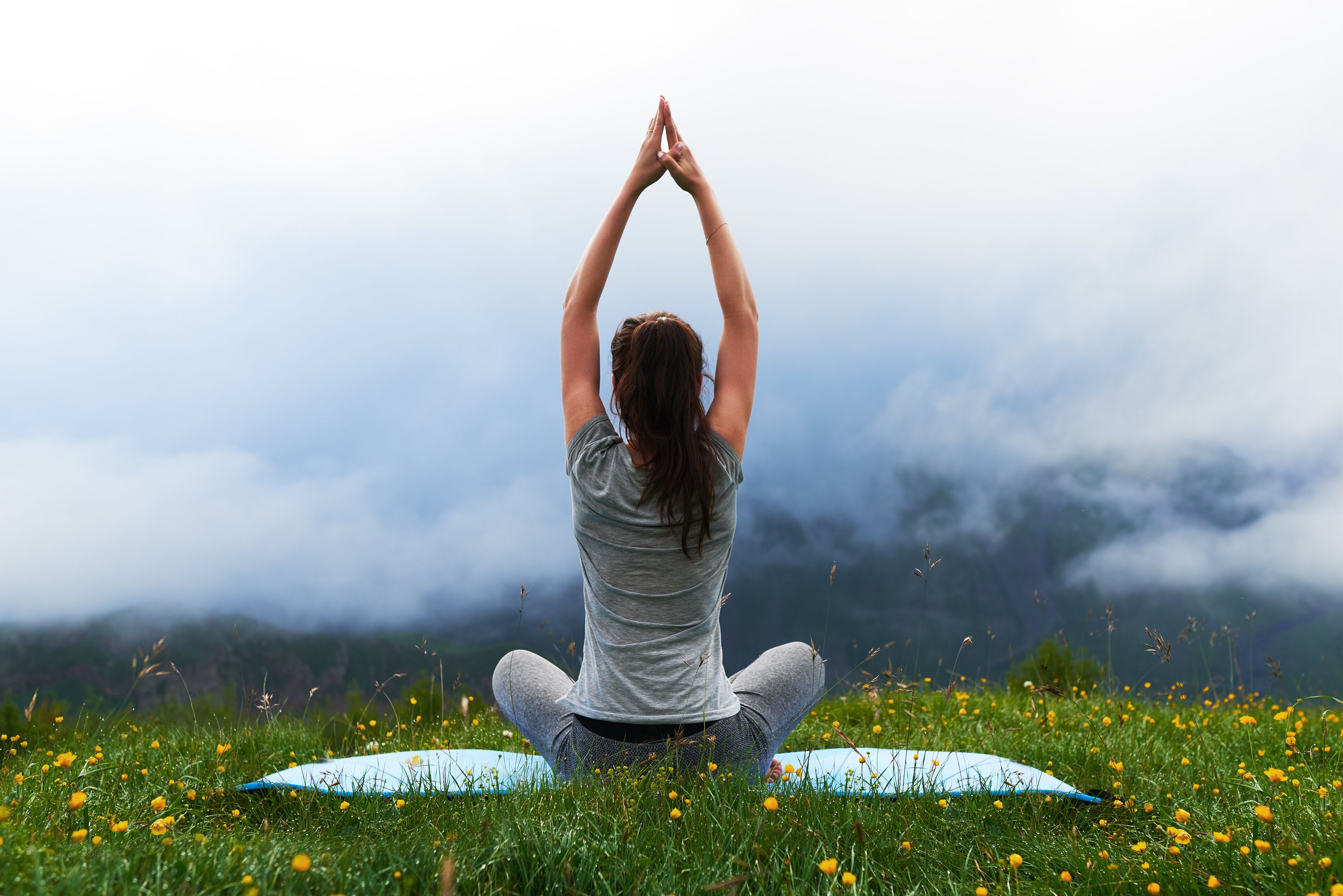 A person sitting cross-legged on a yoga mat in a grassy field with mountains in the background.