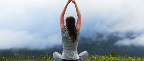 A person sitting cross-legged on a yoga mat in a grassy field with mountains in the background.