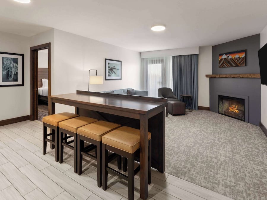 A modern hotel room with a wooden dining table, four bar stools, a flat-screen TV mounted on the wall, a fireplace, and a large window with curtains.