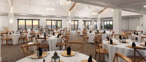 Un gran salón de banquetes con mesas redondas dispuestas para cenar, con manteles blancos y platos de carga dorados.