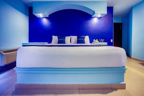 Una cama grande con colchón blanco y ropa de cama azul, una mesita de noche con lámpara, y una lámpara de pared sobre la cama.