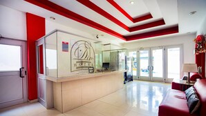 Zona de recepción de un hotel con un separador de vidrio, mostrador de recepción, un sofá rojo y una puerta que da al exterior.