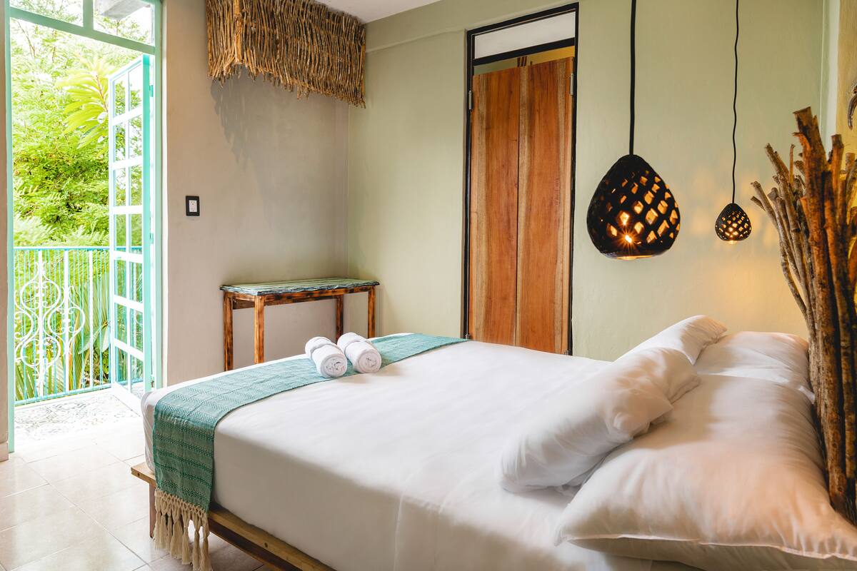 A neatly made bed with white linens and pillows, a wooden nightstand with a green cloth, and a woven wall hanging.