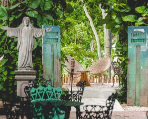 A statue with outstretched arms, a wicker chair, and a bench in a garden setting. -  Straw Hat Tulum (Tulum)