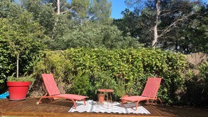 Eine Holzterrasse mit zwei roten Liegestühlen und einem kleinen Tisch, umgeben von dichtem Grün und Bäumen.