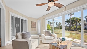 A spacious living room with a large window, a ceiling fan, and a wooden coffee table. - SunnyMar Retreat Beach Access • Heated Pool • Near Historic Bridge Street (Longboat Key)
