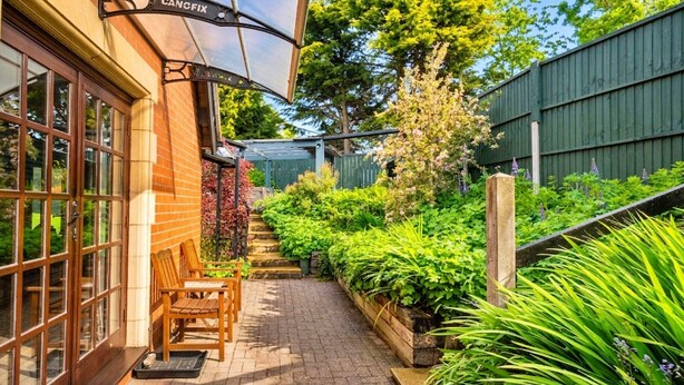 Ein Backsteinhaus mit einer Holzbank und einem Tisch vor der Tür, ein Garten mit verschiedenen Pflanzen und ein Holzzaun.