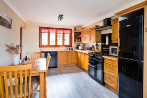 A modern kitchen with wooden cabinets, a black refrigerator, a stove, and a dining area with a wooden table and chairs. - The Grove Annexe (Craven Arms)