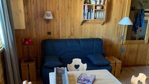 A cozy wooden room with a sofa, a table with chairs, a lamp, and a shelf with books and decorative items.