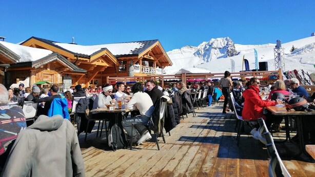 A wooden outdoor seating area with tables and chairs, surrounded by snow-covered mountains and clear blue sky.